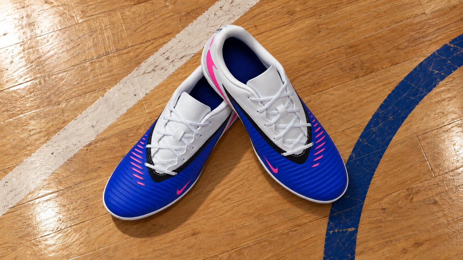 Par de chuteiras de futsal azuis e brancas, com detalhes rosa, sobre piso de madeira de quadra coberta.